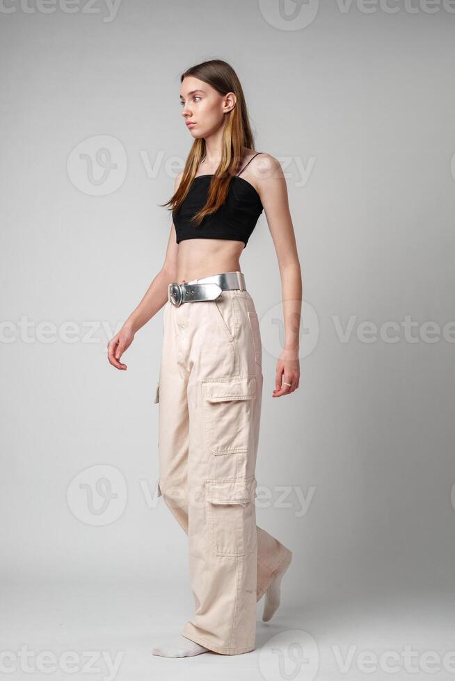 Young Woman Wearing Beige Cargo Pants and a Black Crop Top Standing in Front of a White Wall photo