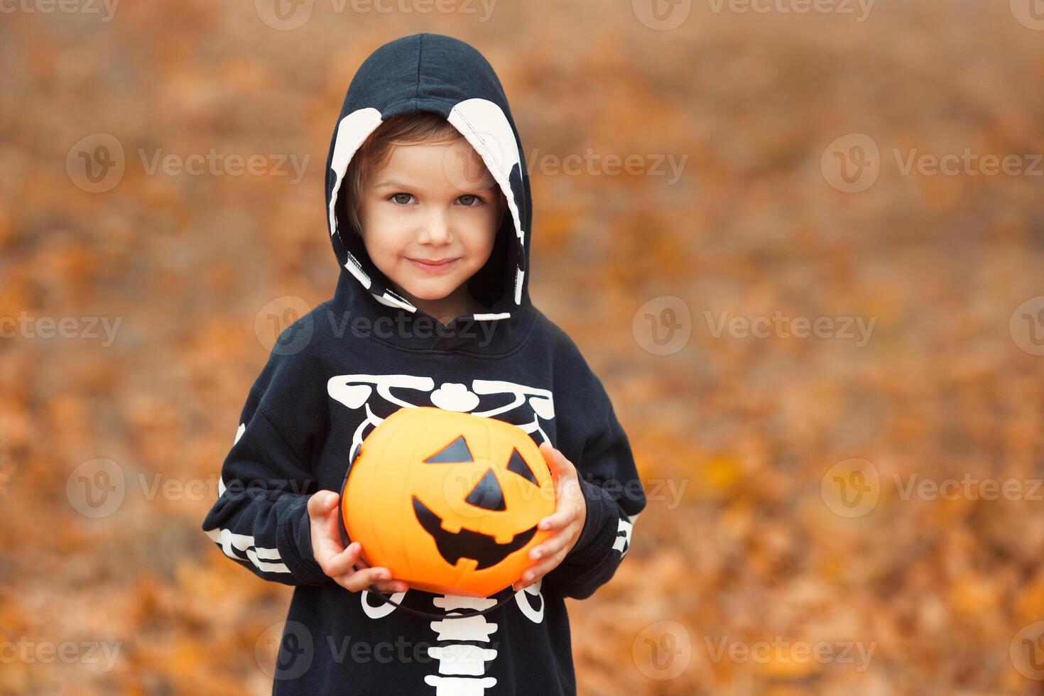 Close-up portrait of a child boy 4-5 years old in a skeleton costume holding a Halloween candy bucket on a background of autumn leaves. Copy space. photo