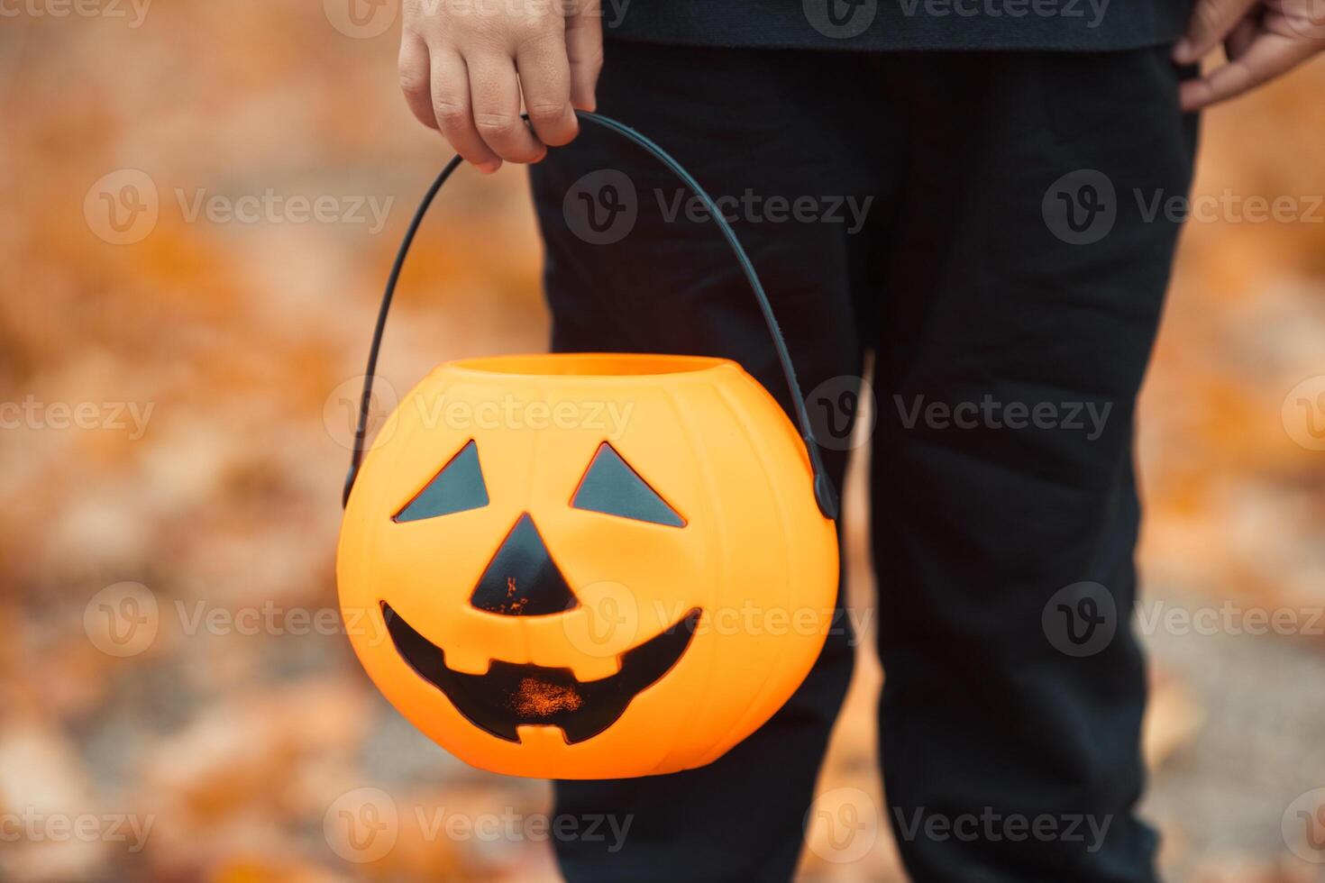 Close-up kid's hand hold a halloween candy bucket in pumpkin shape for trick or treat game. Celebrate all saints day with jack lantern. photo