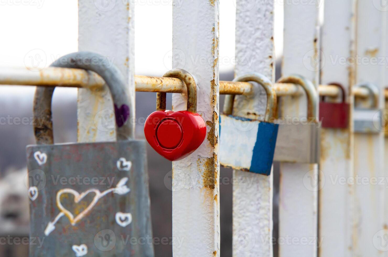 Close-up of various padlocks, including a red heart-shaped lock, attached to a rusty white metal railing symbolizing love, commitment, and memories on a bridge photo