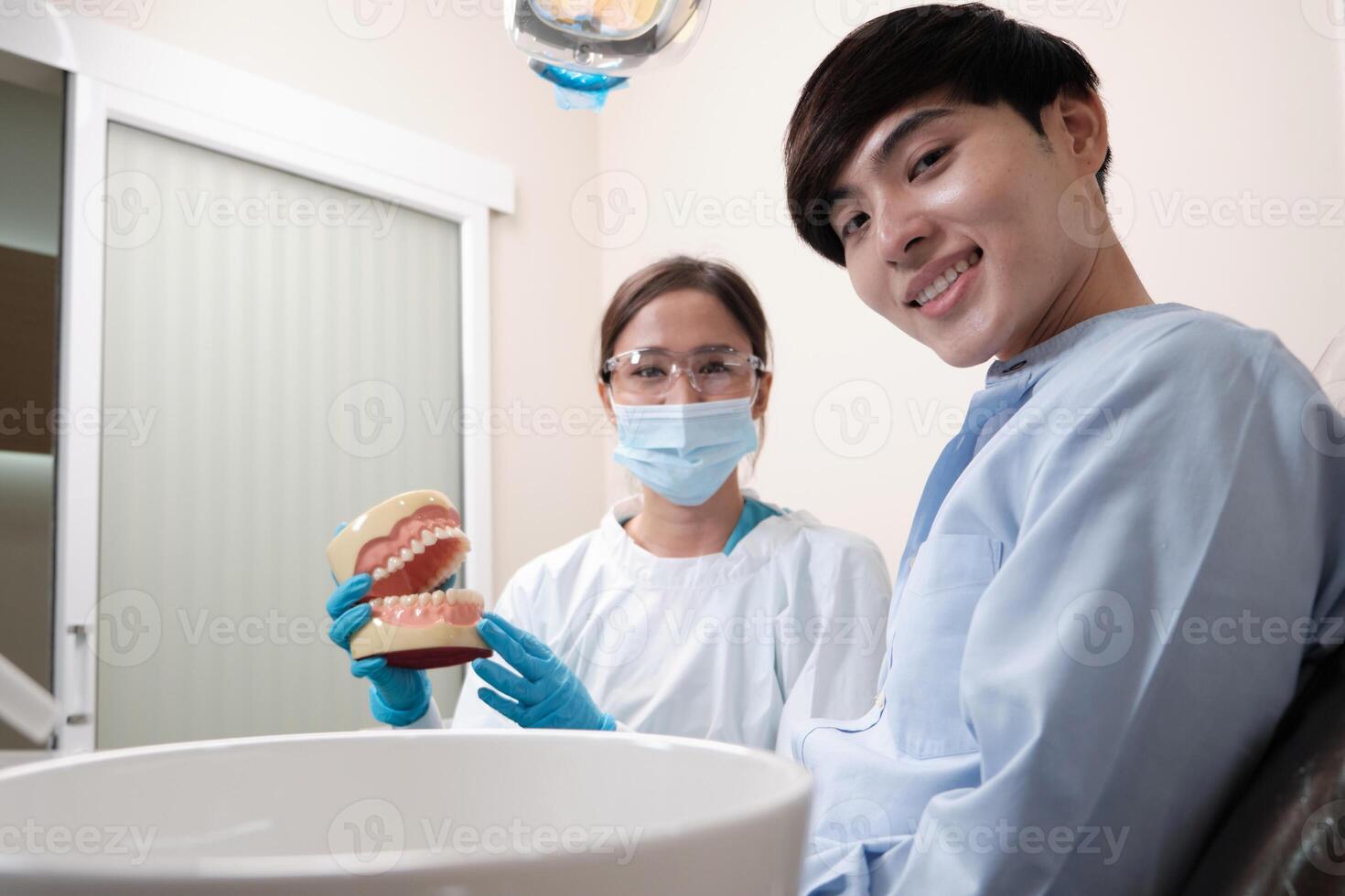 Asian female dentist explaining orthodontic care and whitening to young male patient with teeth model in dental clinic, well-being hygiene, and professional healthcare work in doctor office hospital. photo