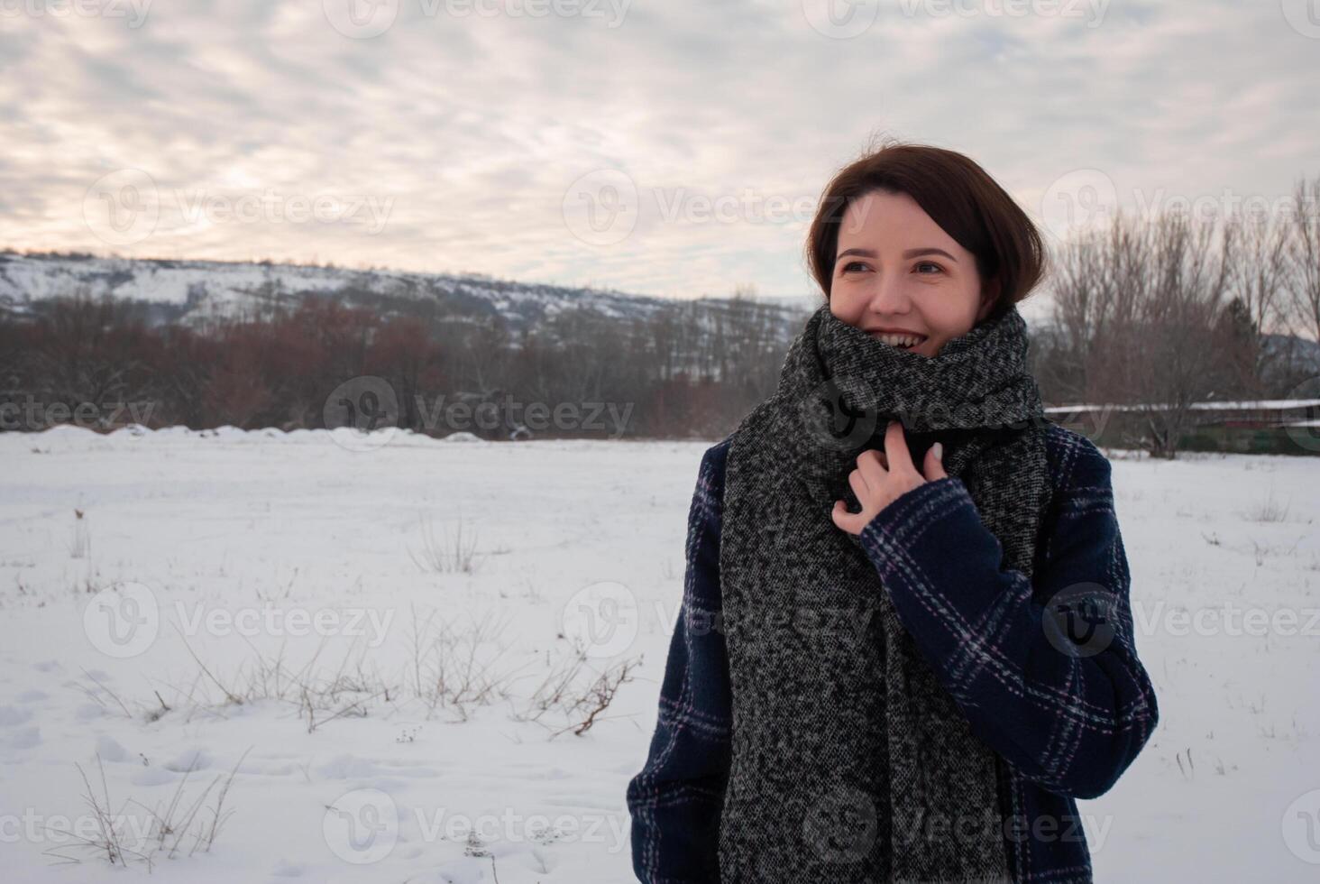 A woman in a dark coat and large scarf smiling in a snowy field, embodying winter coziness and warmth. Highlights comfort and seasonal fashion photo