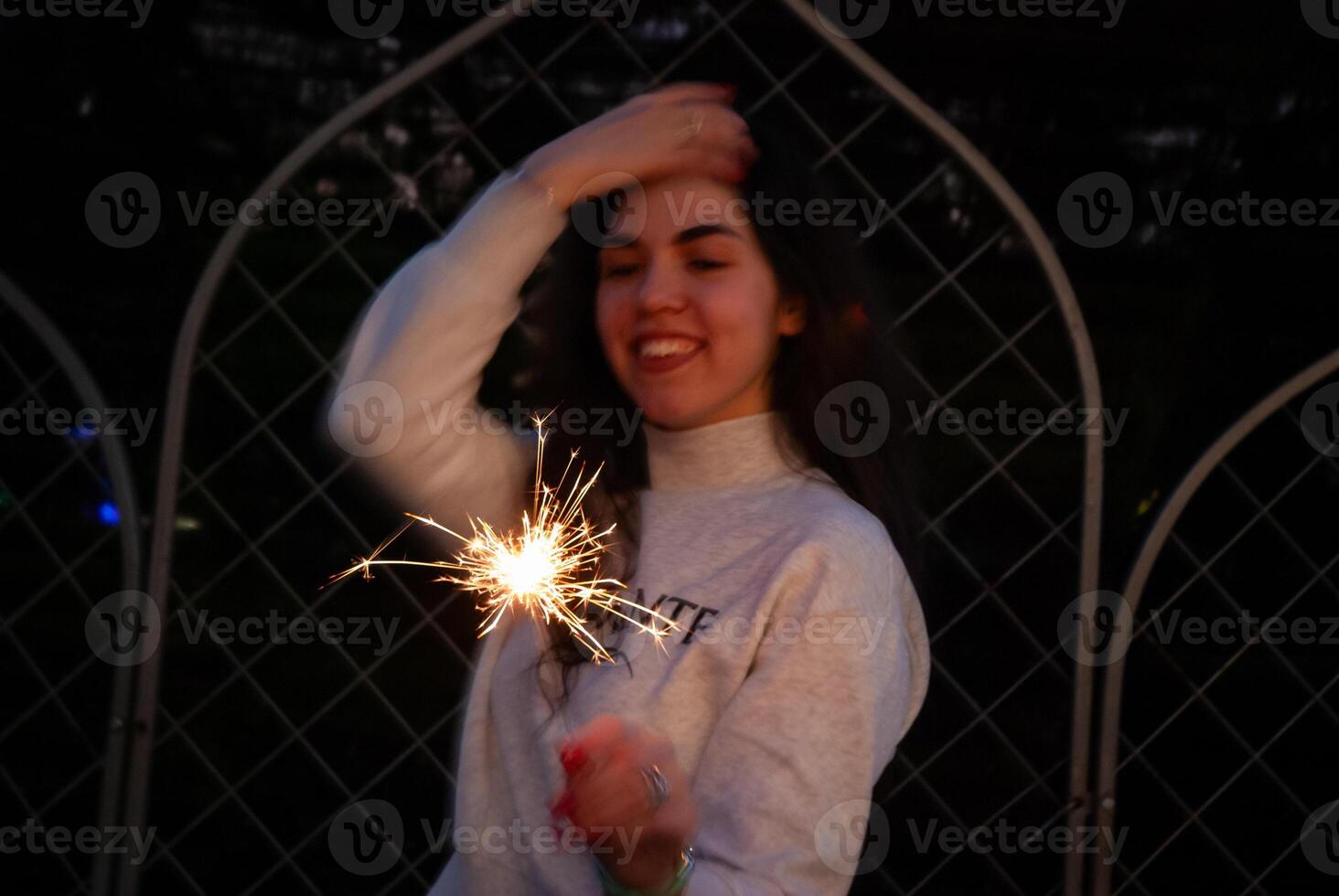 A woman holding a sparkler with a smile in a festive evening setting, green lights in the background adding to the celebratory feel. Warmth and excitement, ideal for holiday promotions photo