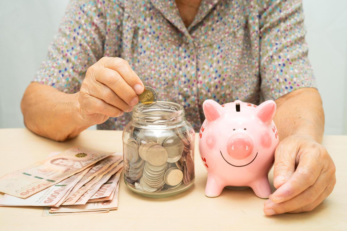 Asian elderly woman putting coin into pink piggy bank for saving money and insurance, poverty, financial problem in retirement. photo