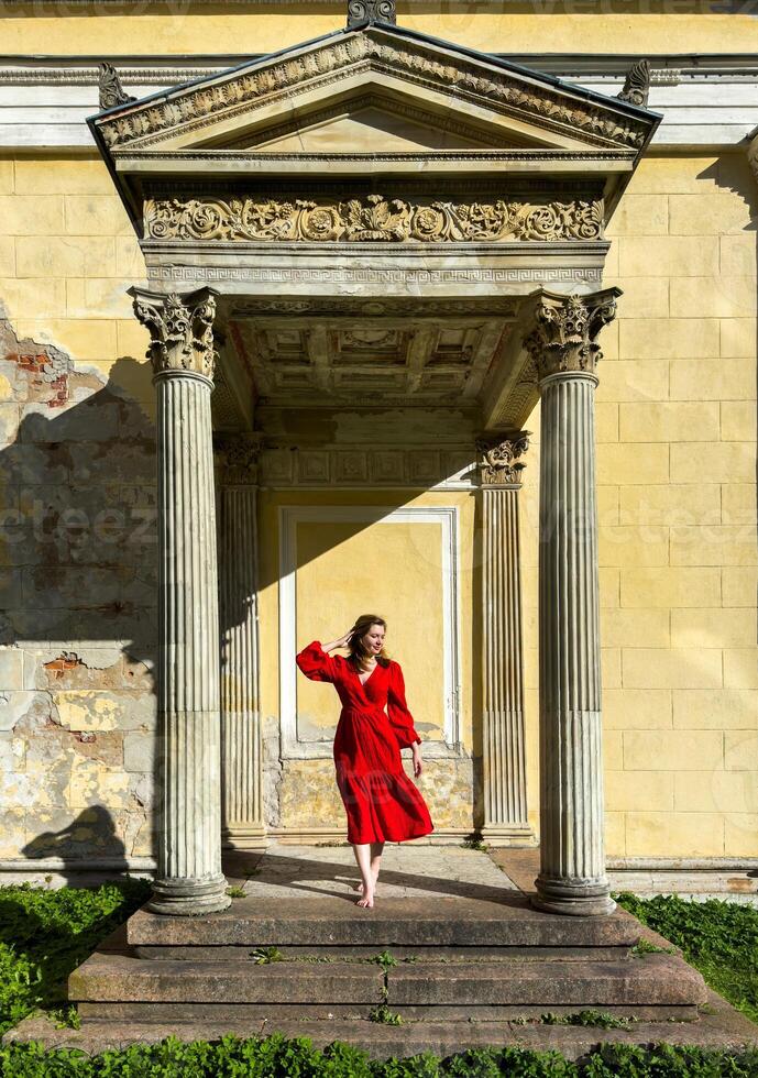 A woman in a red dress stands on the steps of a historic building, framed by classical columns and ornate architecture. The image captures her poised expression and the contrast between her vibrant photo
