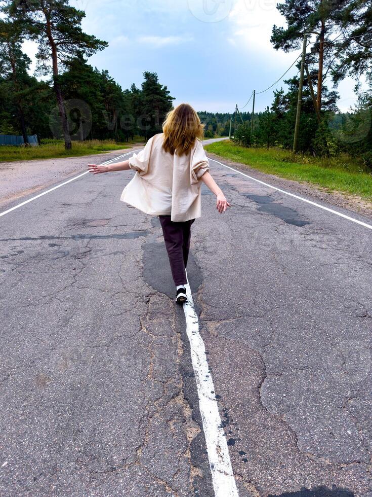 A carefree woman walks down an empty country road lined with pine trees, arms outstretched in the wind. Captured from behind, the scene emphasizes the wide open space, cracked asphalt, and serene photo