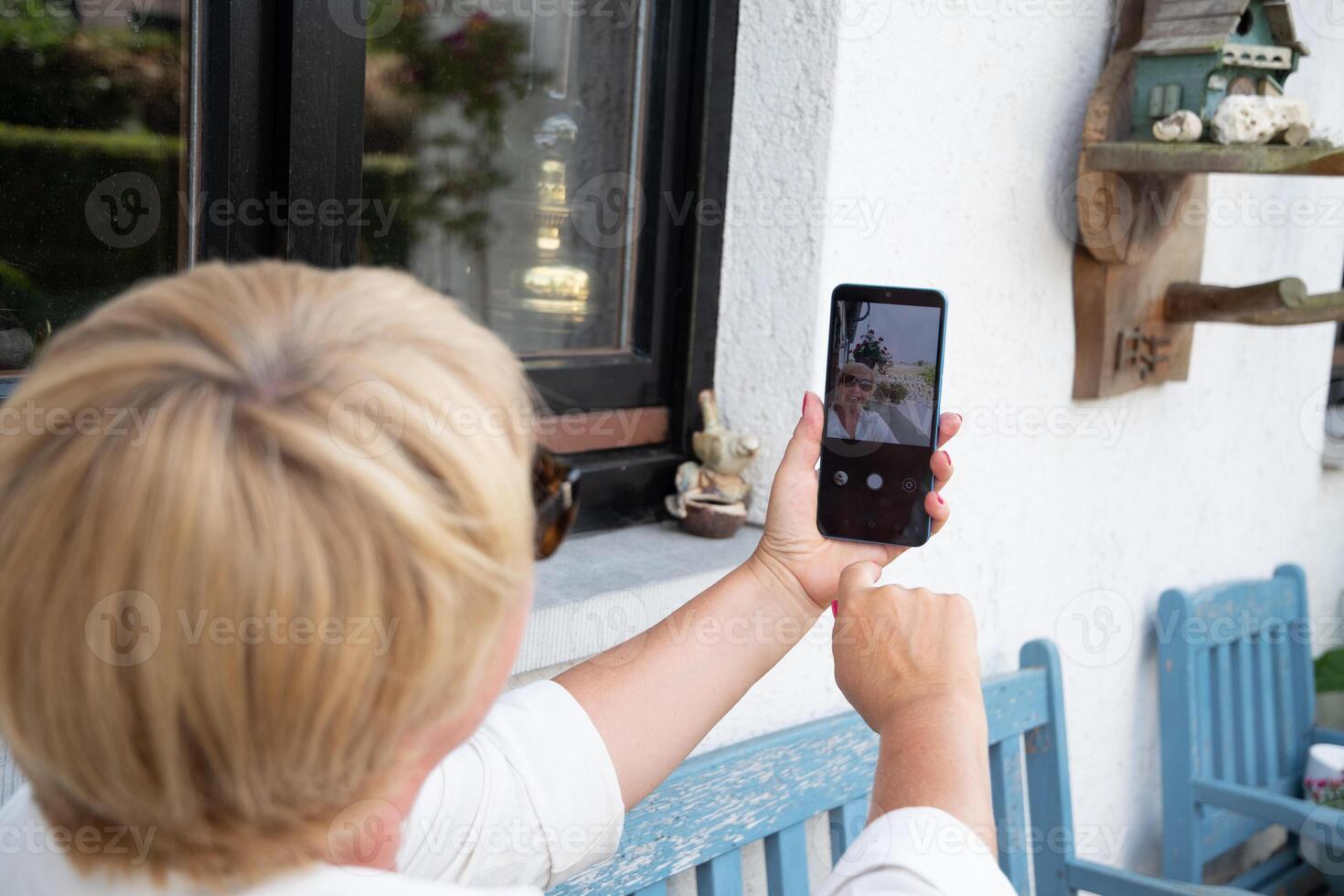Middle aged woman in sunglasses with fashionable short blonde haircut smiling and taking selfie on mobile phone while sitting on blue bench on terrace with flowers, photo
