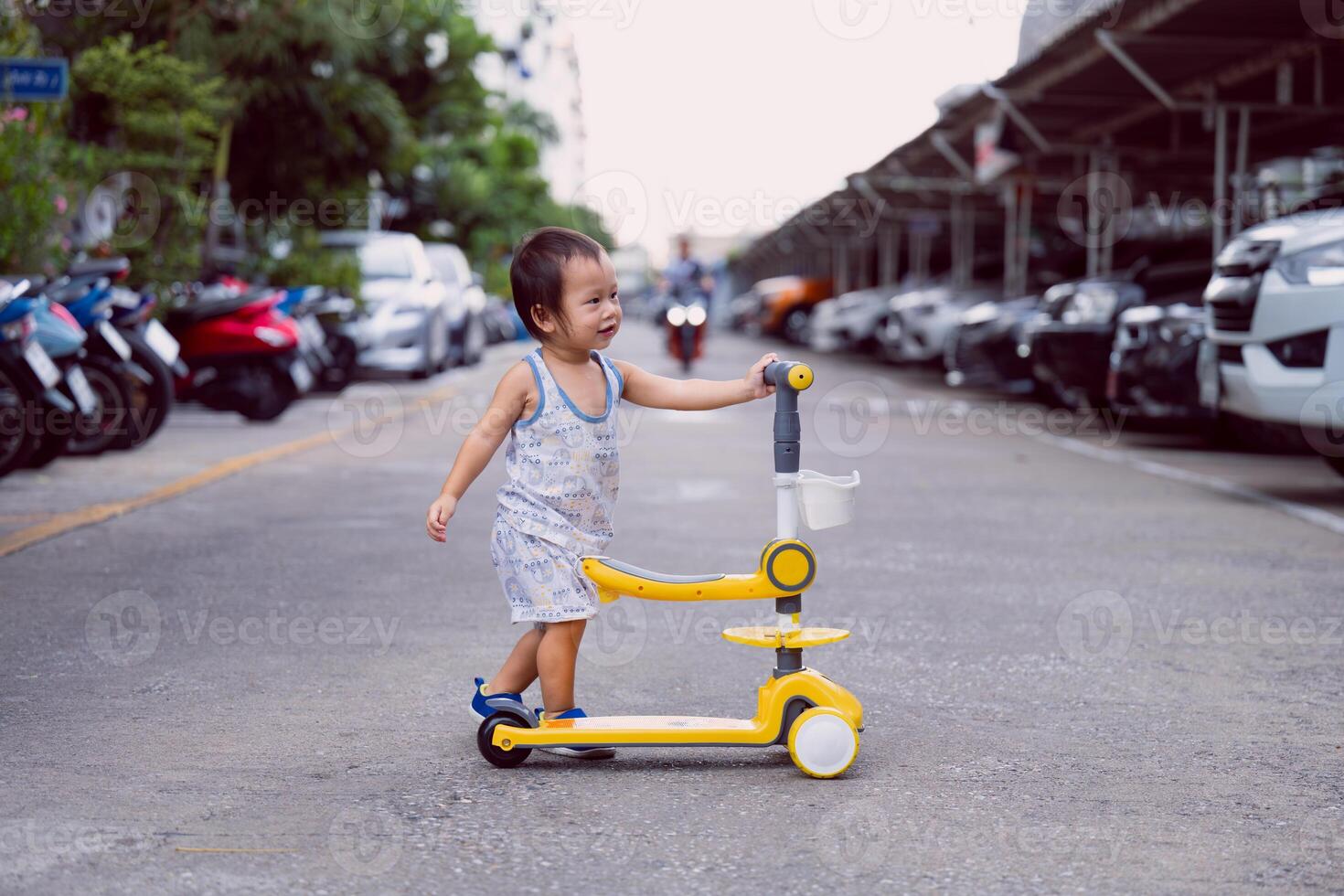 Asian Child Boy is playing with a yellow scooter on a village street. In the background of the image there is a motorcycle driving straight ahead. Concept of little kid and road safety. photo