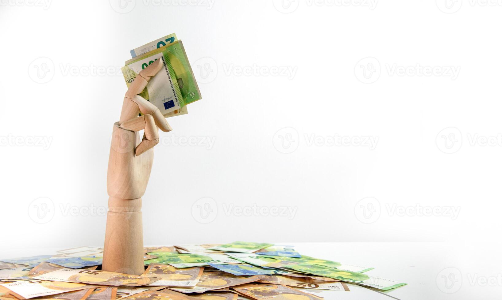 Wooden hand with Euro bills emerges from money pile against white background. Hand in foreground holds bills high, with pile below photo