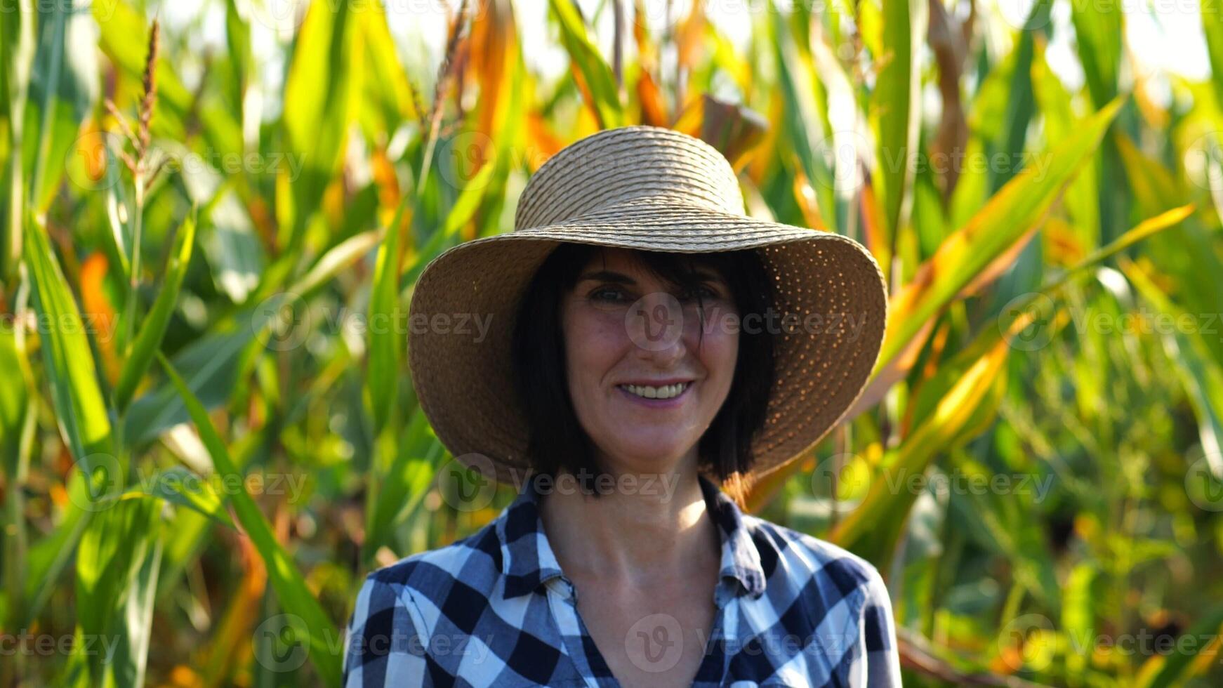 Happy smiling female farmer looks into camera standing near corn field. Portrait of adult beautiful agronomist in straw hat with maize meadow at background. Agricultural business concept. Close up photo