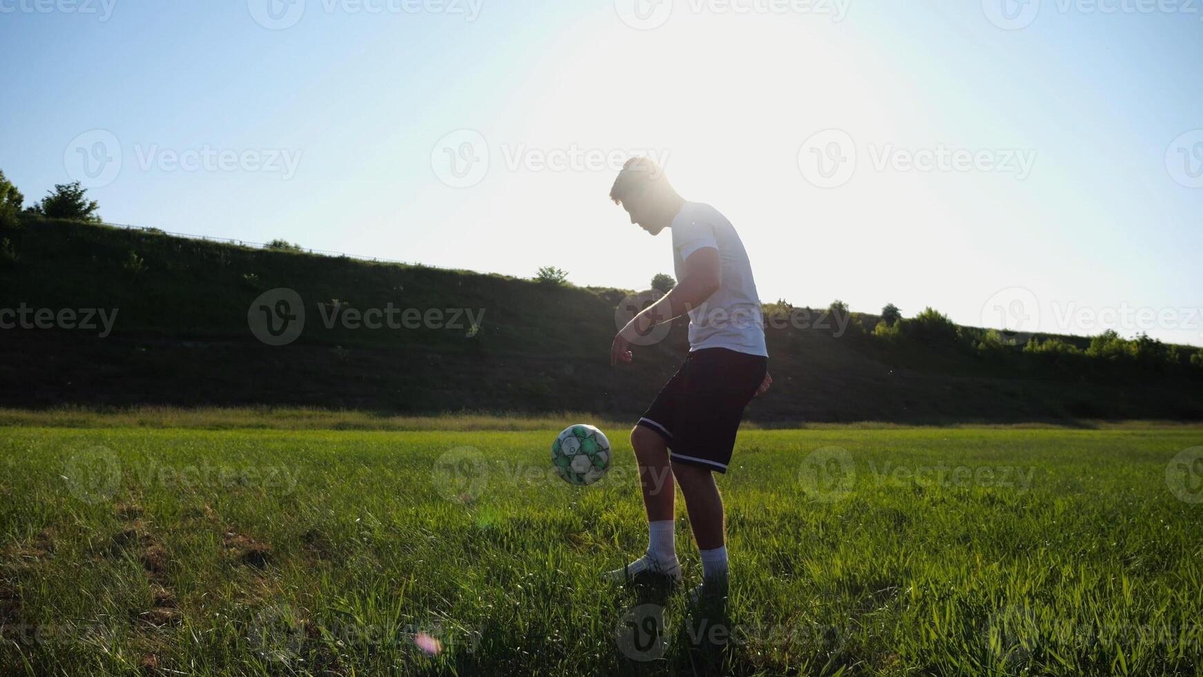 Sportsman juggling soccer ball on stadium at sunny day. Young man kicking ball at green field. Professional footballer practicing tricks at meadow with sunlight at background. Freestyle football. photo