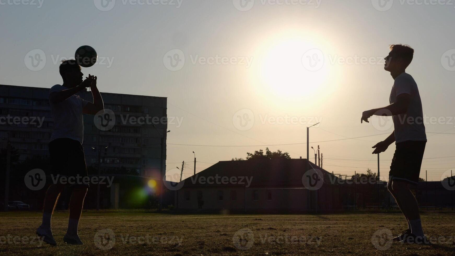 Silhouette of professional footballers kicking soccer ball with their heads on stadium at sunset. Two sportsmen showing tricks with ball while passing it to each other at field. Freestyle football photo