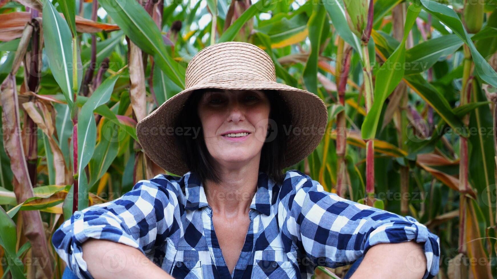 Happy smiling female farmer looks into camera sitting near corn field. Portrait of adult tired agronomist in straw hat with maize meadow at background. Concept of agricultural business. Close up photo