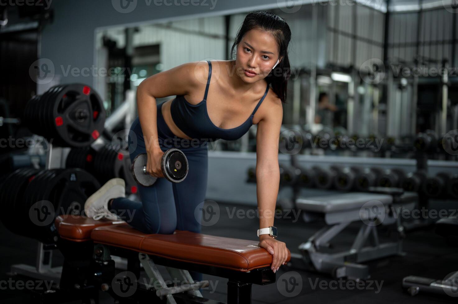A fit Asian woman is performing dumbbell tricep kickbacks on a bench press, training at a gym. photo