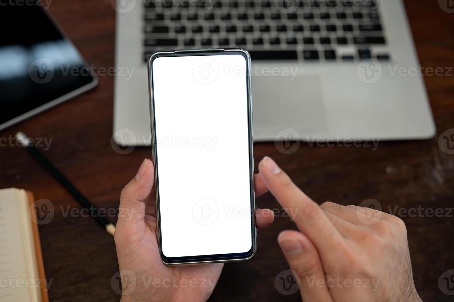 A top view of a man holding a smartphone with a white screen mockup above a desk. photo