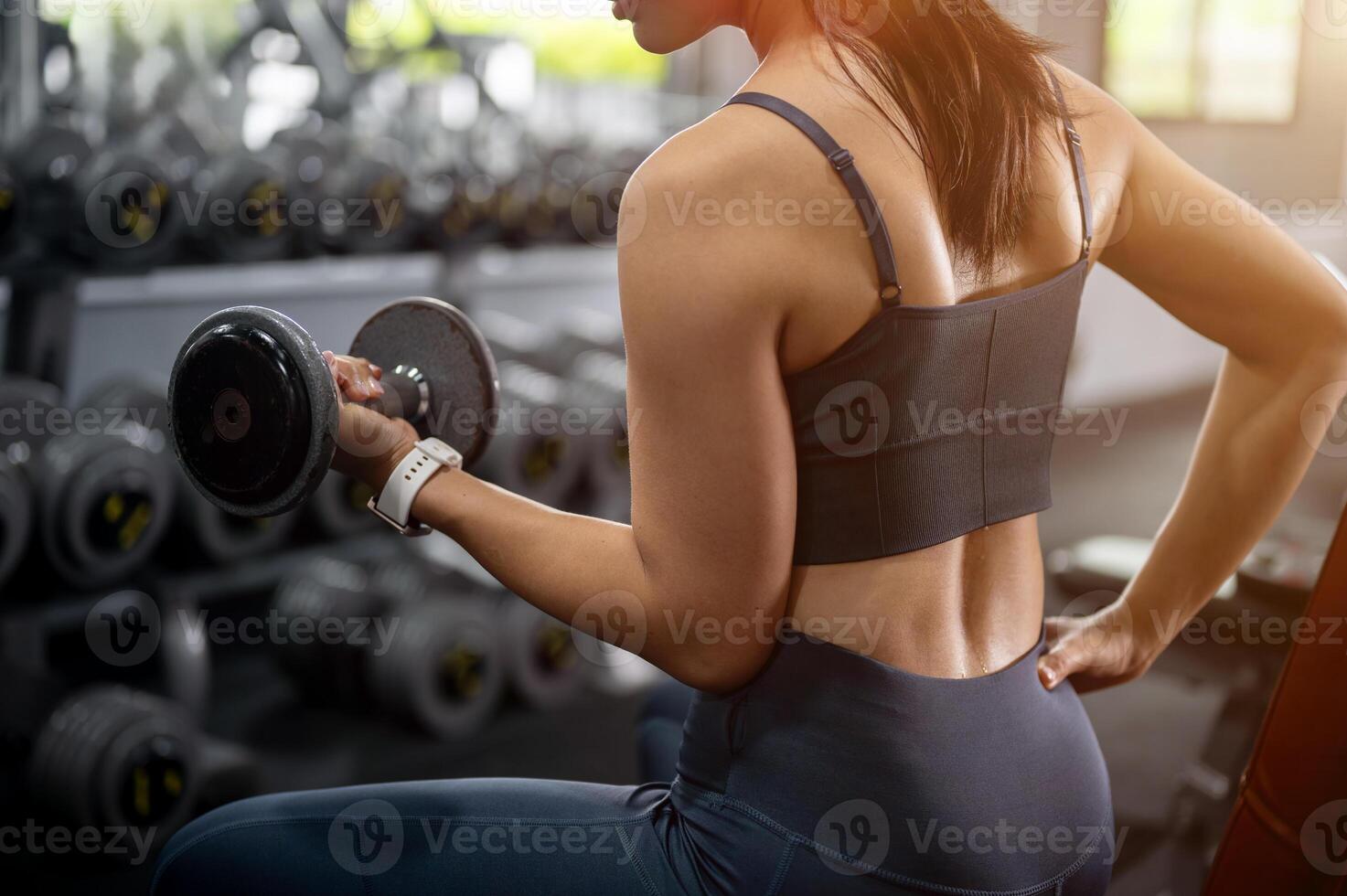 A sweaty, sporty Asian woman in athletic wear sitting on a bench press and lifting a dumbbell. photo