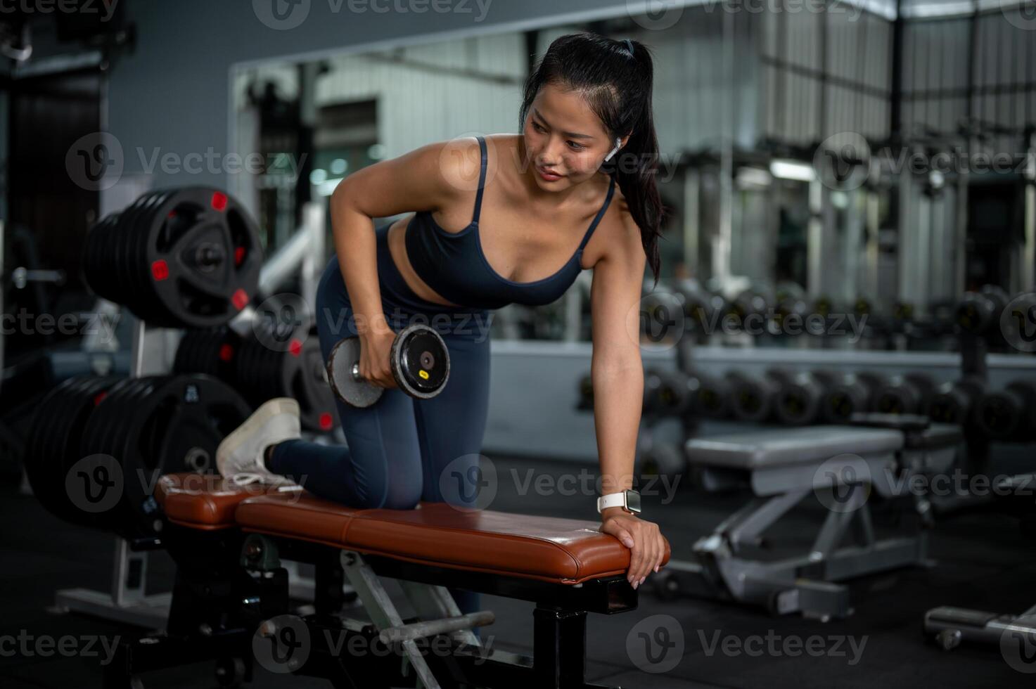 A fit Asian woman is performing dumbbell tricep kickbacks on a bench press, training at a gym. photo