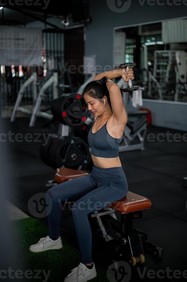 A sporty Asian woman is sitting on a bench press and performing a dumbbell overhead tricep extension photo