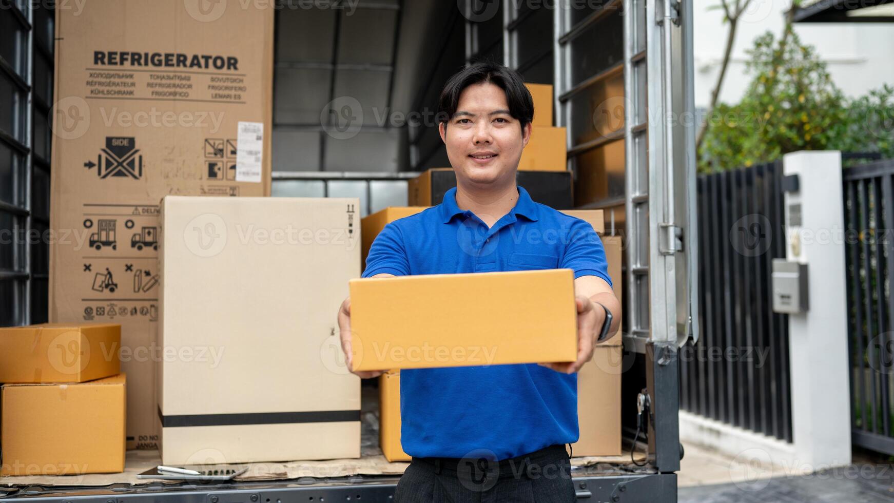 A smiling Asian delivery guy standing at the back of his truck, carrying a package for delivery. photo