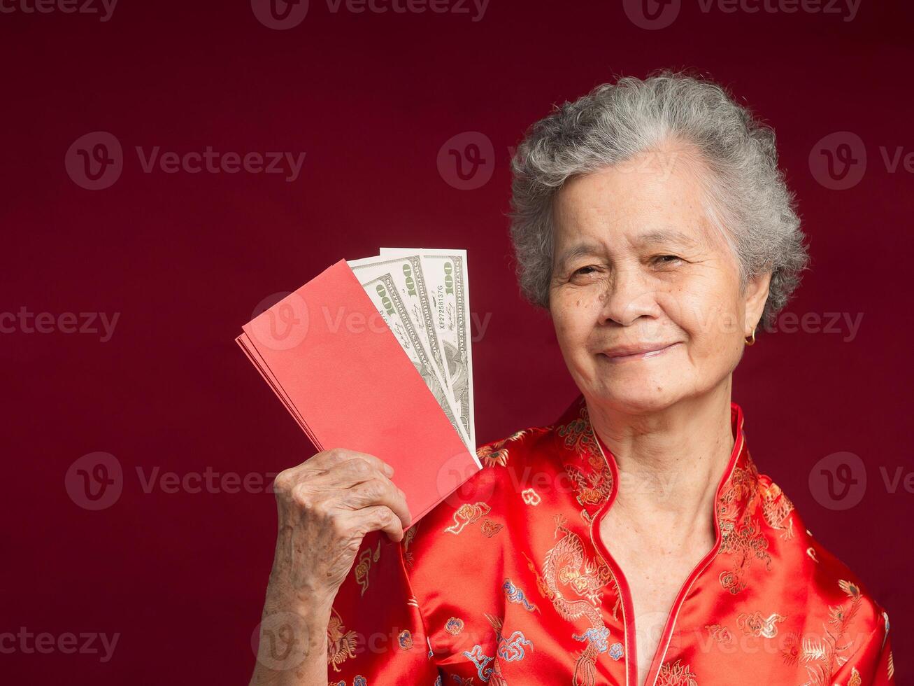 Senior woman wearing a traditional cheongsam qipao dress holding Angpao with US dollar banknote while standing over a red background photo