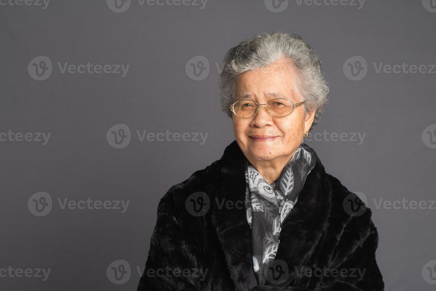 Senior Asian woman in a sweater and scarf looking at the camera with a smile while standing on a gray background photo
