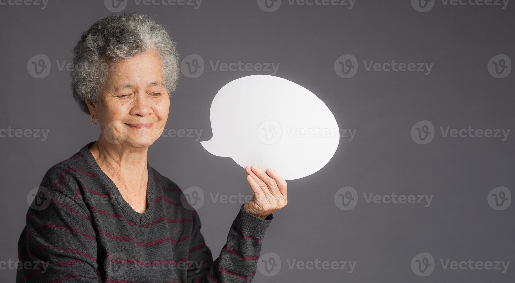 A speech bubble concept. A senior woman holding and looking at a blank bubble balloon while standing on a gray background photo
