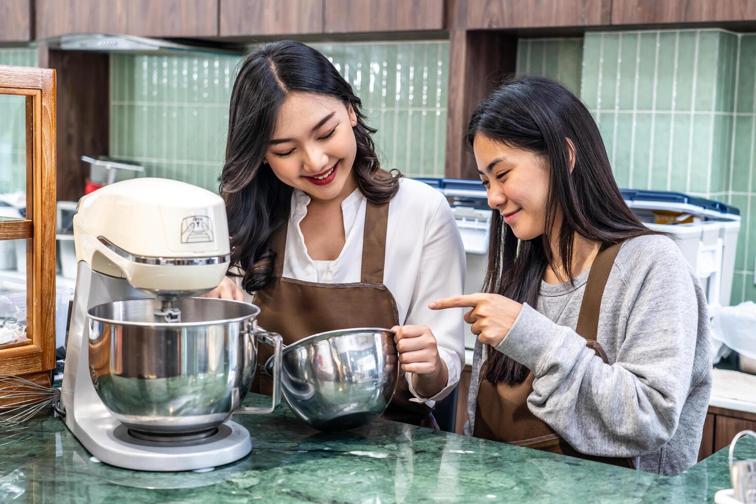 Two young asian women friends and aprons making cream for cupcakes, Using electric mixer, Preparing Cookies, Cooking, Homemade, Made specifically to sell photo
