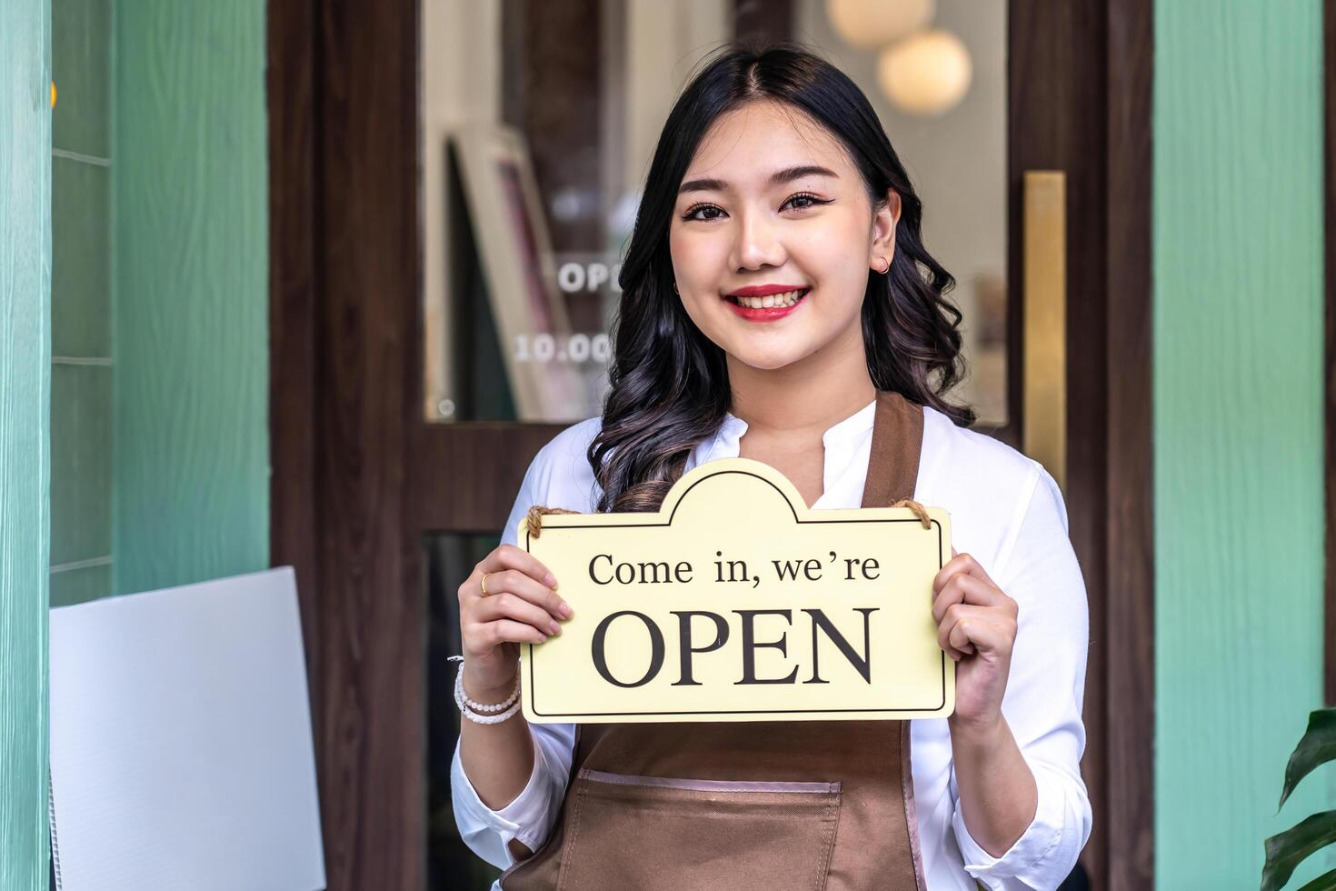 Business owner happy beautiful young asian woman in apron looking at camera, we're open sign on front door smiling welcoming clients to new cafe, People and start-up concept. photo
