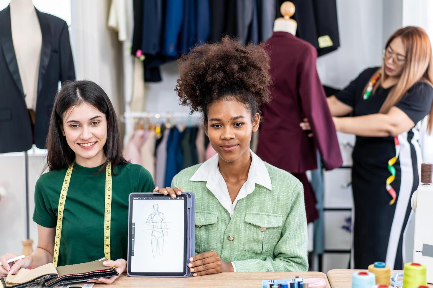 Young two fashion designers use digital tablet meeting with graphic designer discuss show sample dress design new collection on screen on desk while working in sewing workshop photo