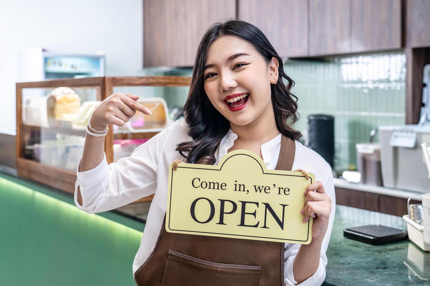 Happy beautiful Asian female waitress or younger shopkeeper in an apron holding open sign while standing at cafe or in restaurant, with pleasured smiling, welcome, business owner startup ideas photo
