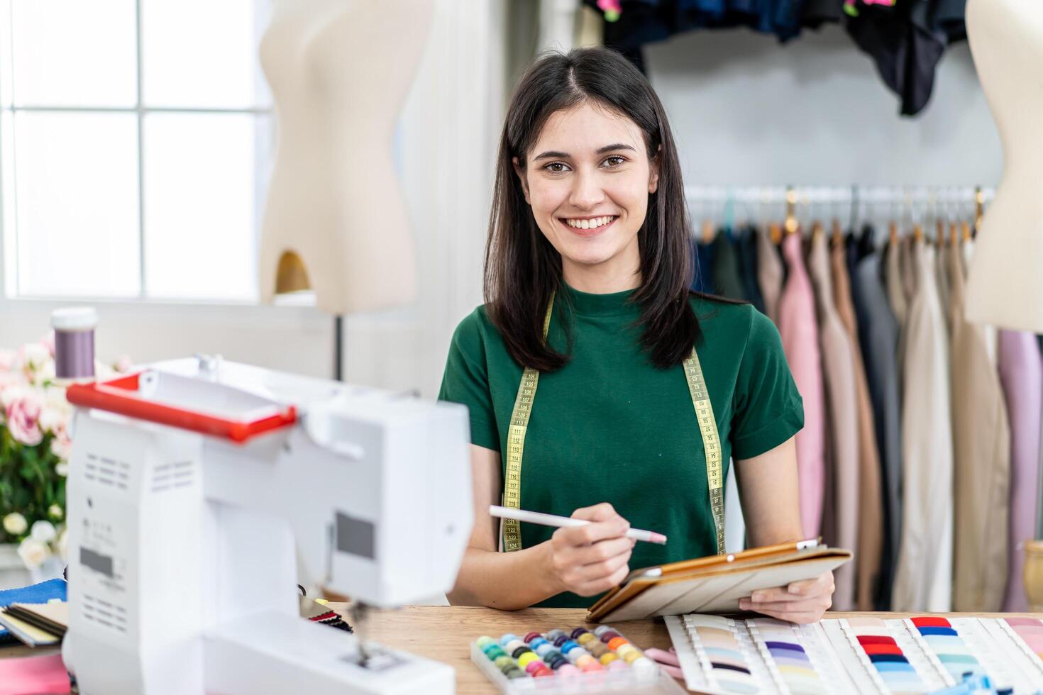 Smiling caucasian female fashion designer works in studio by idea drawing sketches with digital tablet and colorful fabric for a dress design collection, choose clothing colors for tailoring photo