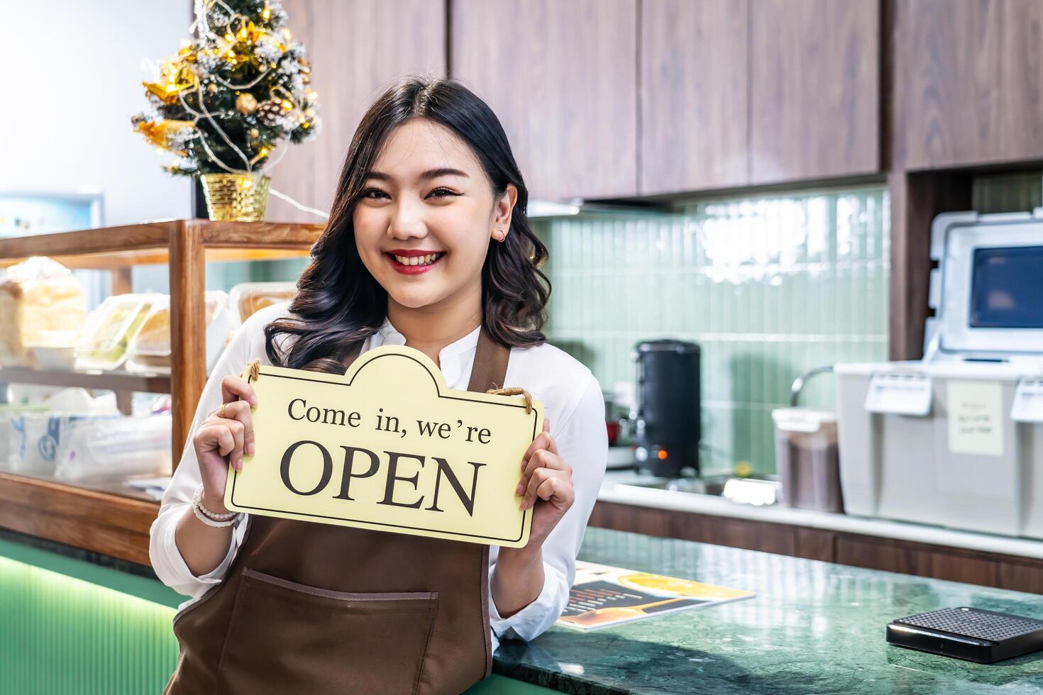 Happy beautiful Asian female waitress or younger shopkeeper in an apron holding open sign while standing at cafe or in restaurant, with pleasured smiling, welcome, business owner startup ideas photo