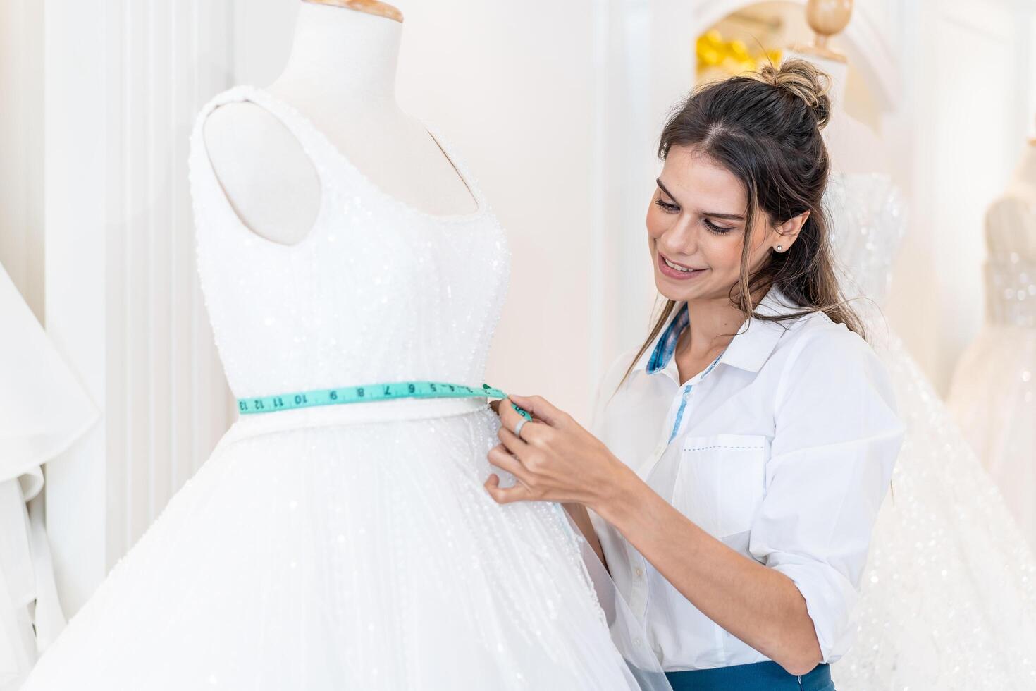 Smiling caucasian designer woman or is bridal shop owner with tape measure measures wedding dress on mannequin in workshop, Small business entrepreneur wedding planner and tailor designer concept photo