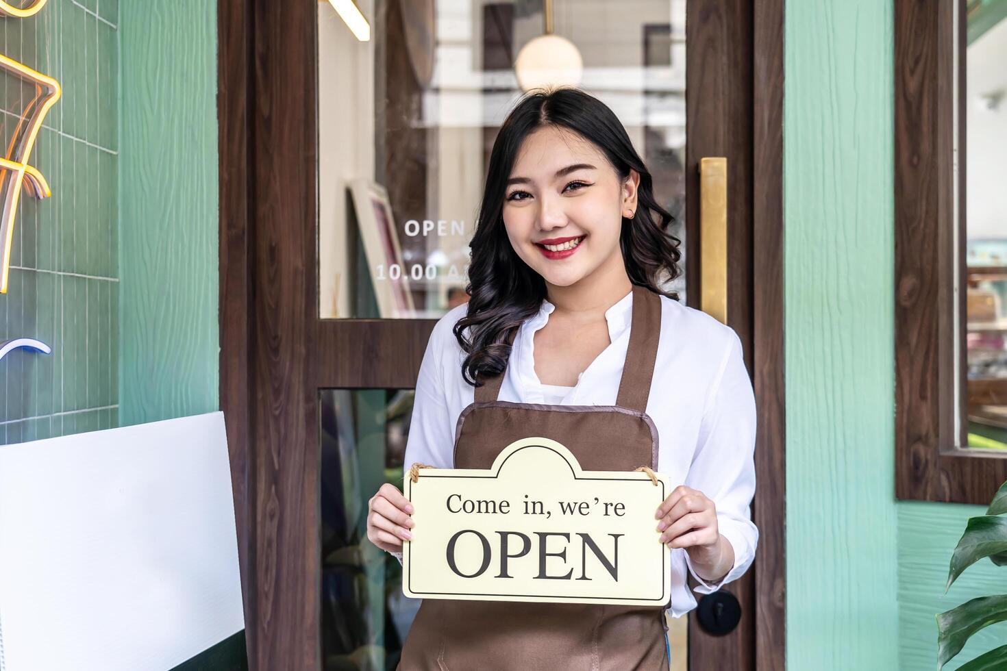 Business owner happy beautiful young asian woman in apron looking at camera, we're open sign on front door smiling welcoming clients to new cafe, People and start-up concept. photo