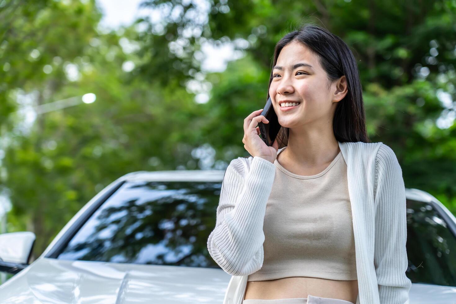Beautiful young asian girl tourist calling phone standing near car at summer weekend. Asian woman talking at smartphone on the road with trees Female enjoys talking phone photo