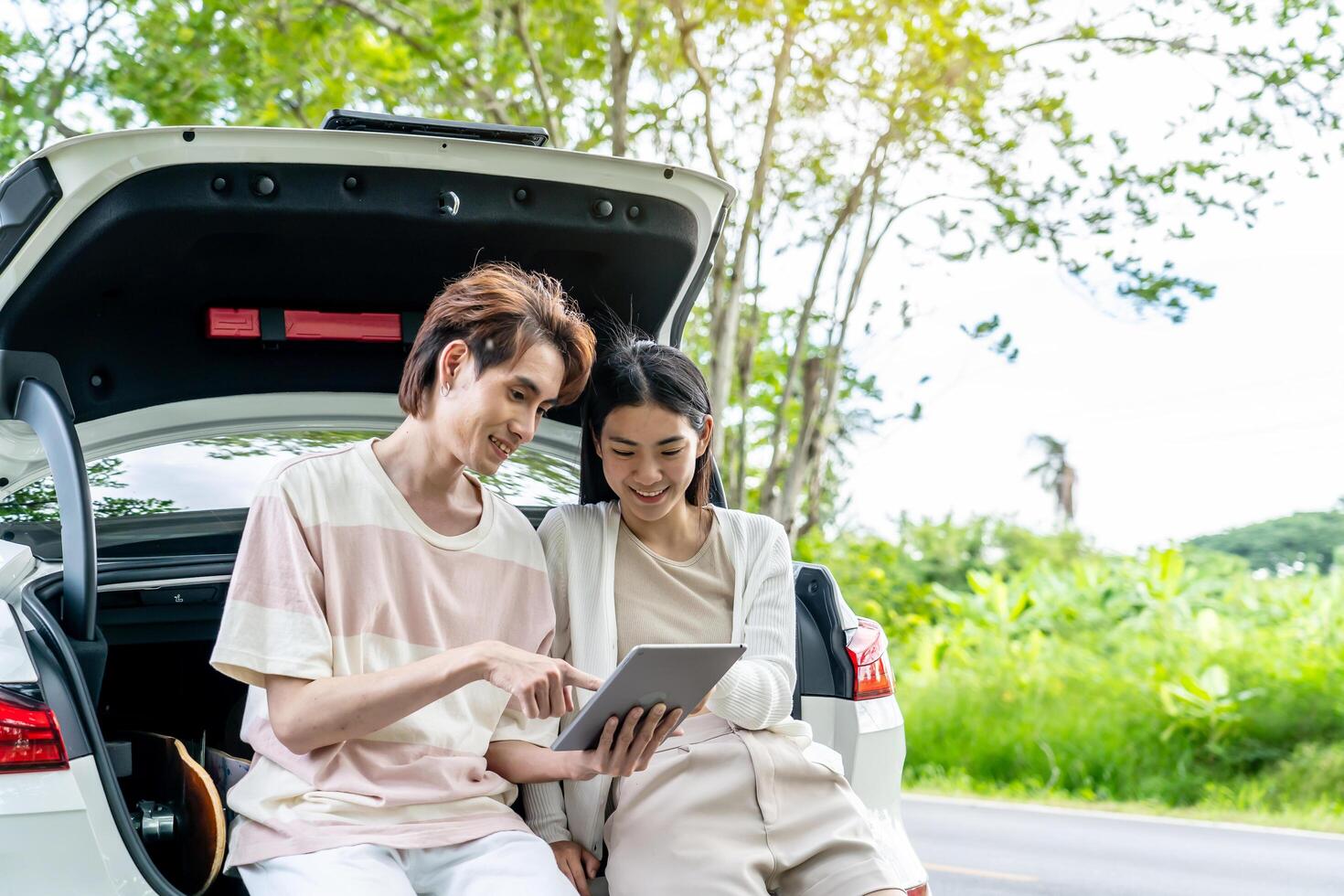 Traveler couple on road trip, man and woman sitting together in car trunk using map in tablet during the trip, Two young boy and girl travel, Two friend traveling on vacation, searching road in map photo