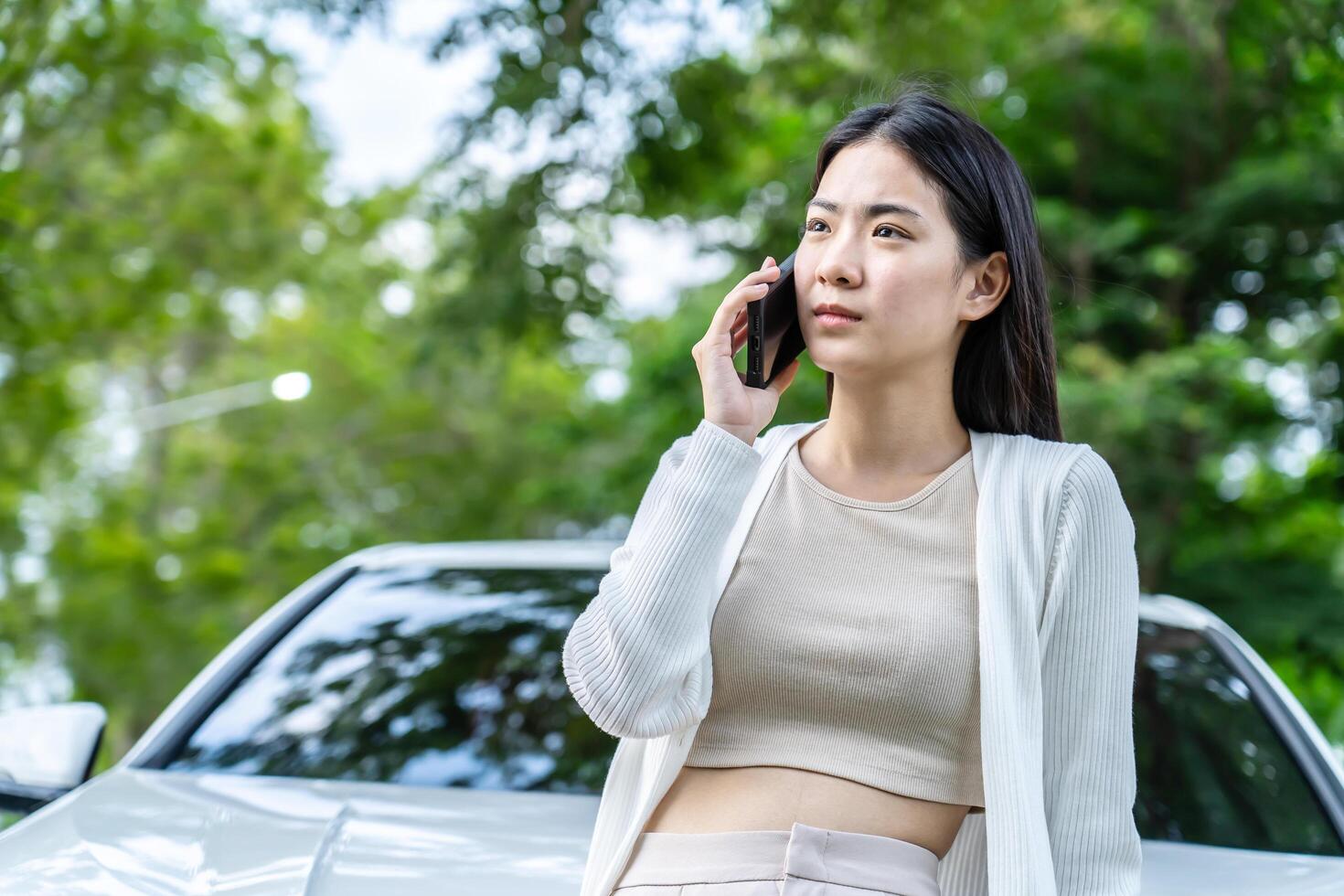 Anxious young asian woman standing near a broken car with popped up hood calling talking on phone. Portrait of worried lady alone with damaged vehicle outdoors photo