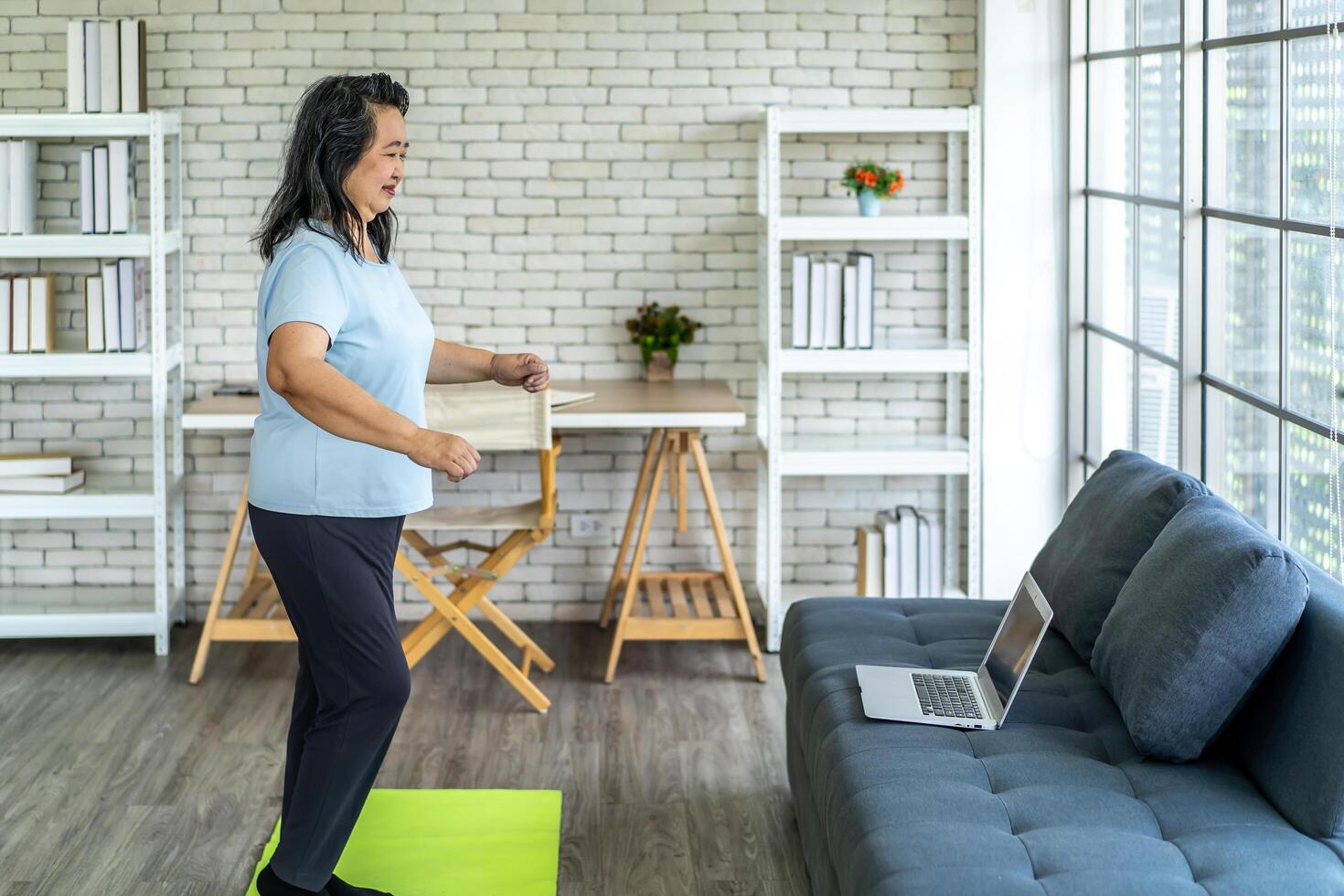 Elderly asian woman watching tutorial doing stretching fitness exercise yoga. Elderly women training online courses on laptop while exercising in living room. Workout training online concept photo