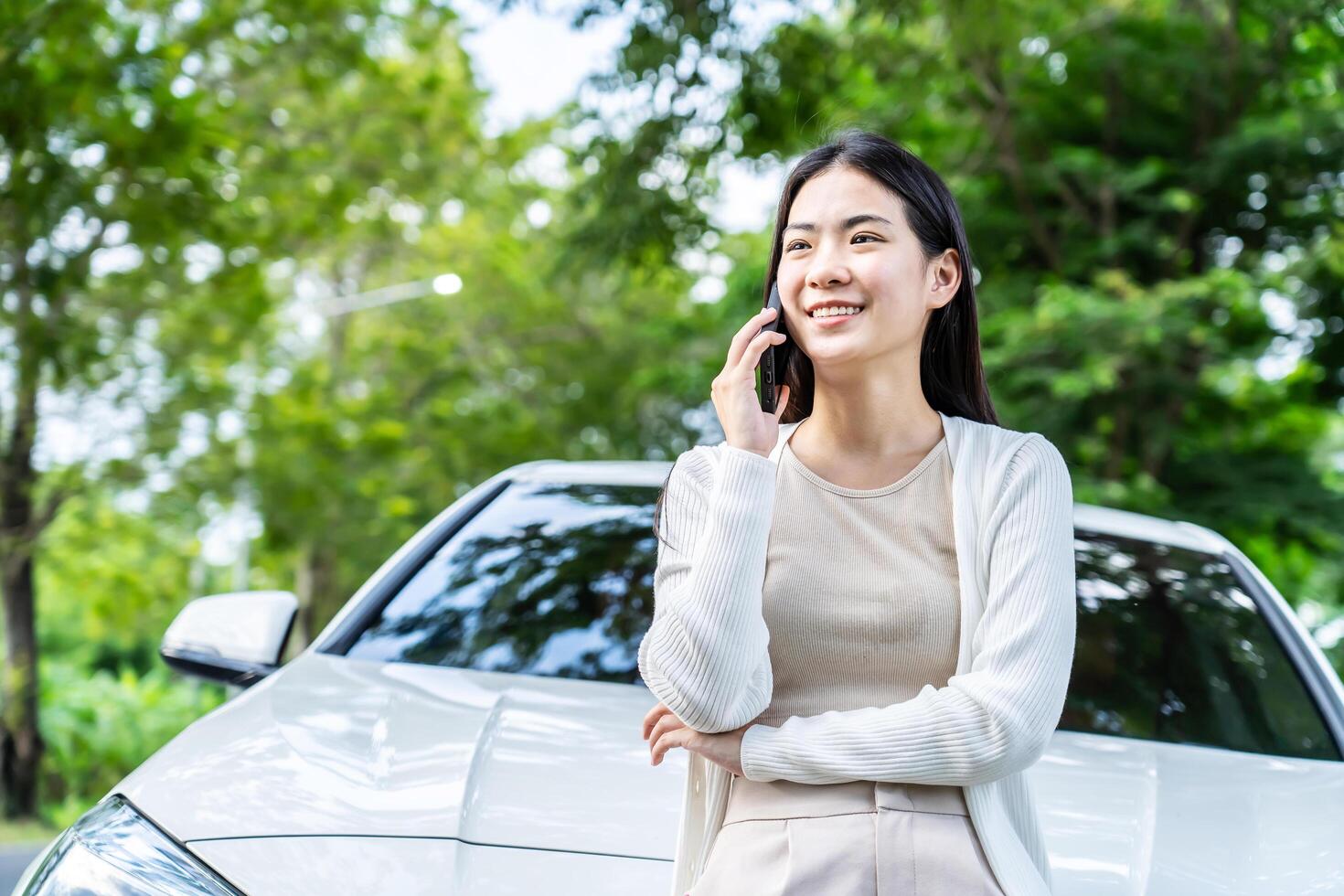 Beautiful young asian girl tourist calling phone standing near car at summer weekend. Asian woman talking at smartphone on the road with trees Female enjoys talking phone photo
