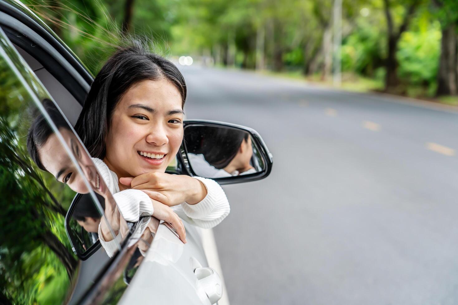 Happy cute asian woman enjoying the breeze sitting in a car looking out of the window car while having a road trip watching a beautiful sunset tree view, Driving road trip on vacation photo