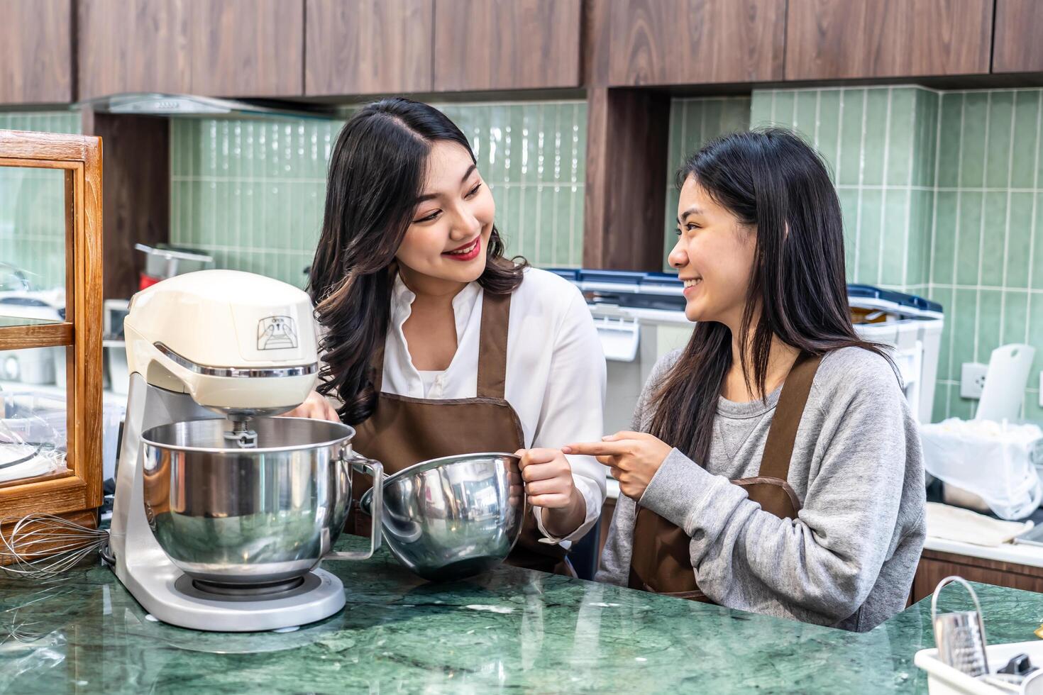 Two young asian women friends and aprons making cream for cupcakes, Using electric mixer, Preparing Cookies, Cooking, Homemade, Made specifically to sell photo