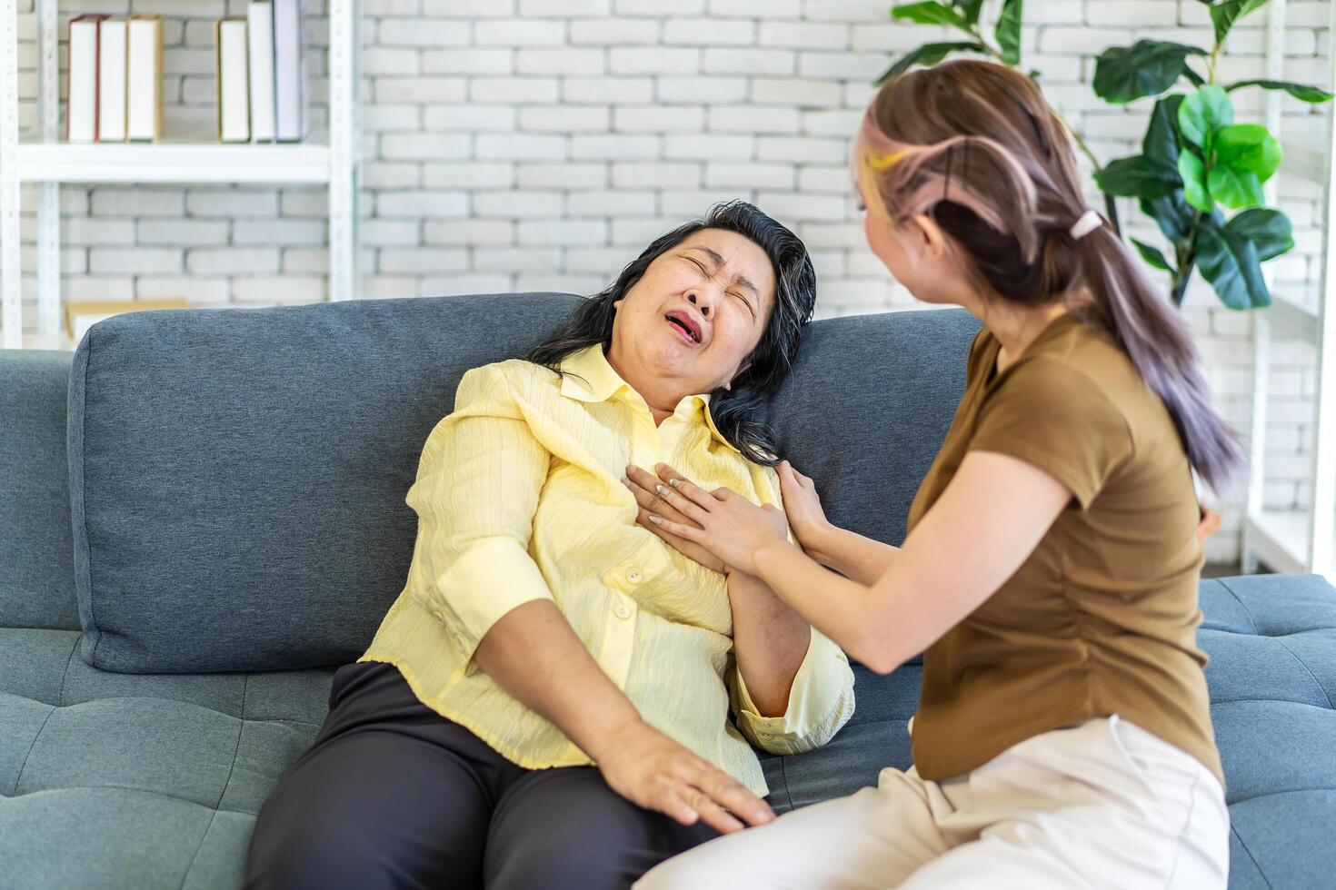 Elderly asian woman having chest pain or heart attack, clutching and having difficult breathing, Loving daughter take care of elderly mother while having left chest pain on sofa. photo