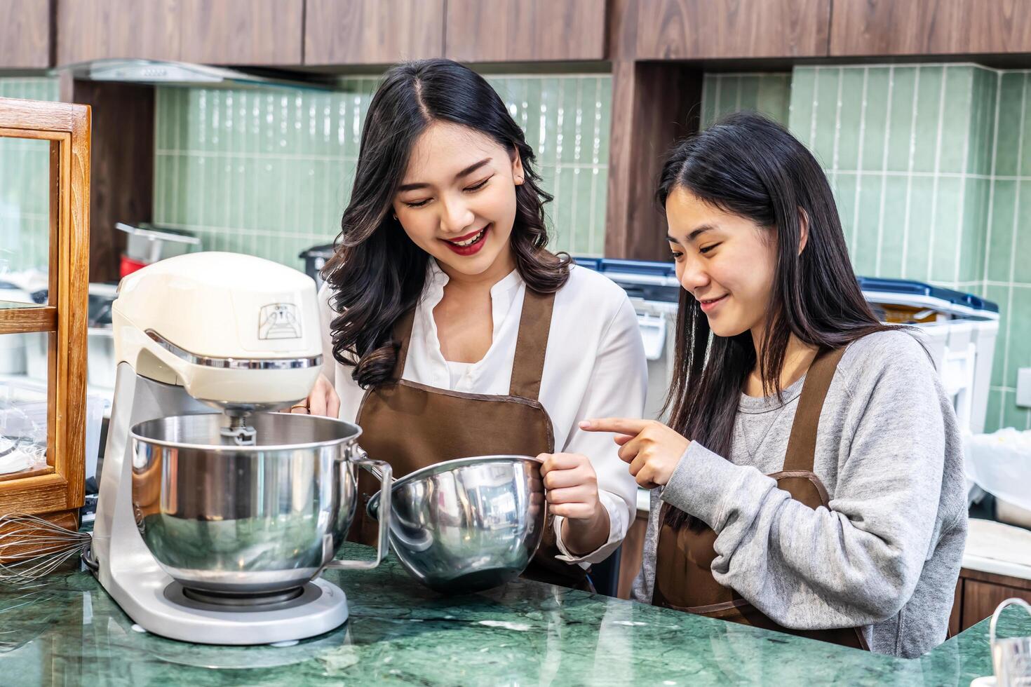 Two young asian women friends and aprons making cream for cupcakes, Using electric mixer, Preparing Cookies, Cooking, Homemade, Made specifically to sell photo