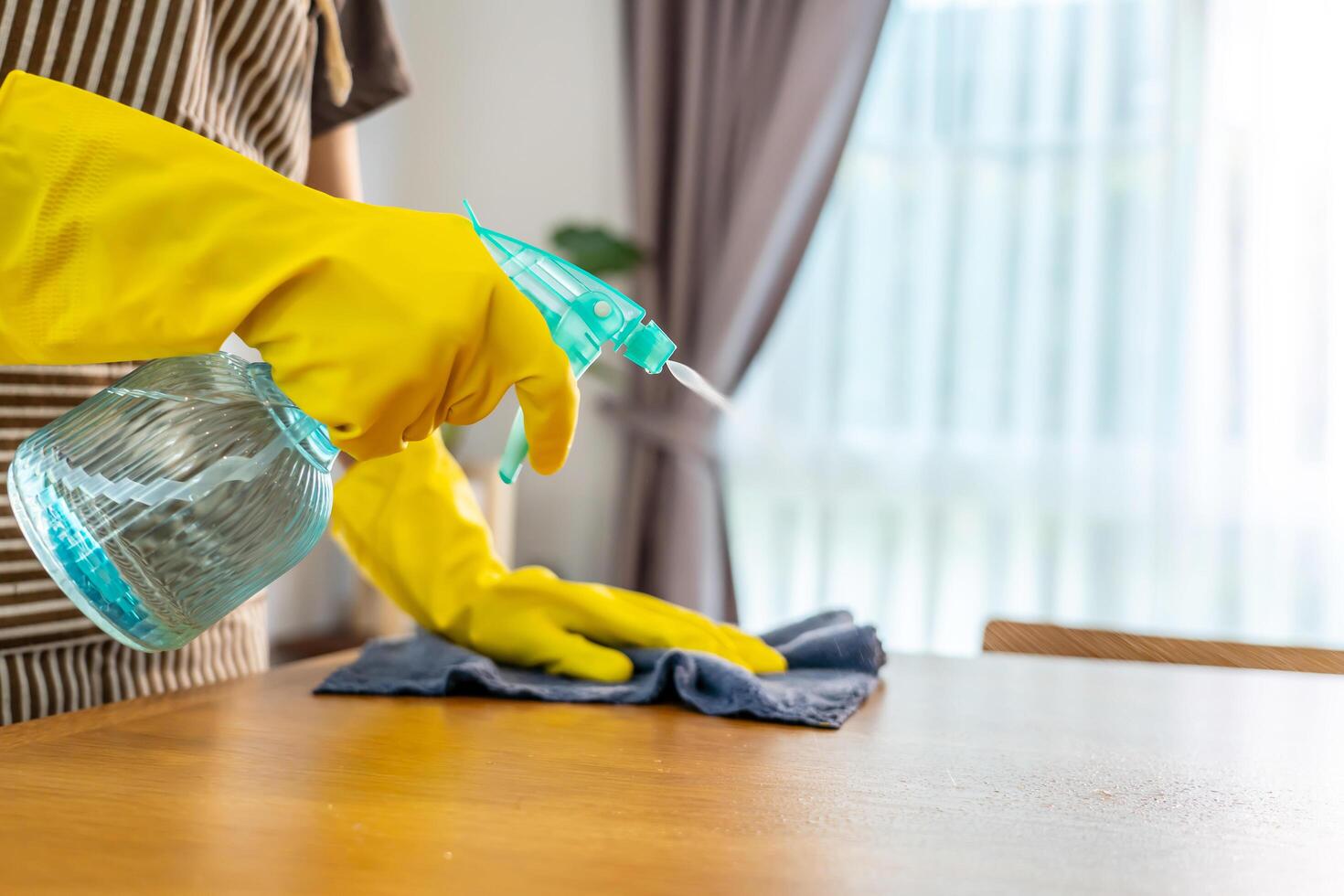 Close-up of hand woman in gloves and apron polishing or wiping wooden table, detergent and chemical bottle for dust, bacteria and disinfection, furniture surface and housekeeping maintenance at home photo