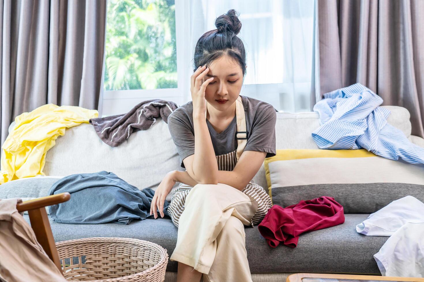 Stressed young woman asian sitting on the sofa in the living room and among a mess at home, woman sitting among scattered clothes on sofa and ignoring, woman frowning and letting out soft sigh. photo