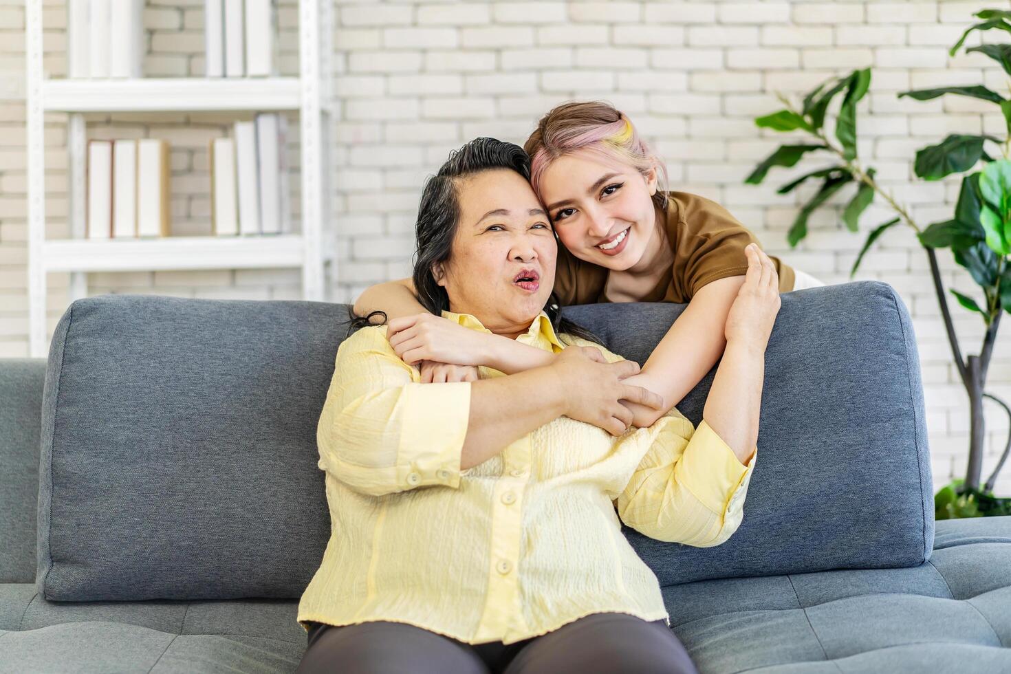 Happy asian young daughter embracing from back senior mother in law feeling respect and love, mature mom and adult daughter hugging and talking with looking at tv, spending leisure time together photo