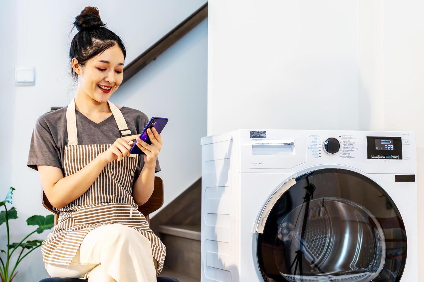 Young asian woman are playing with mobile phone in home sitting near the washing machine, while waiting for the washing machine to finish working photo