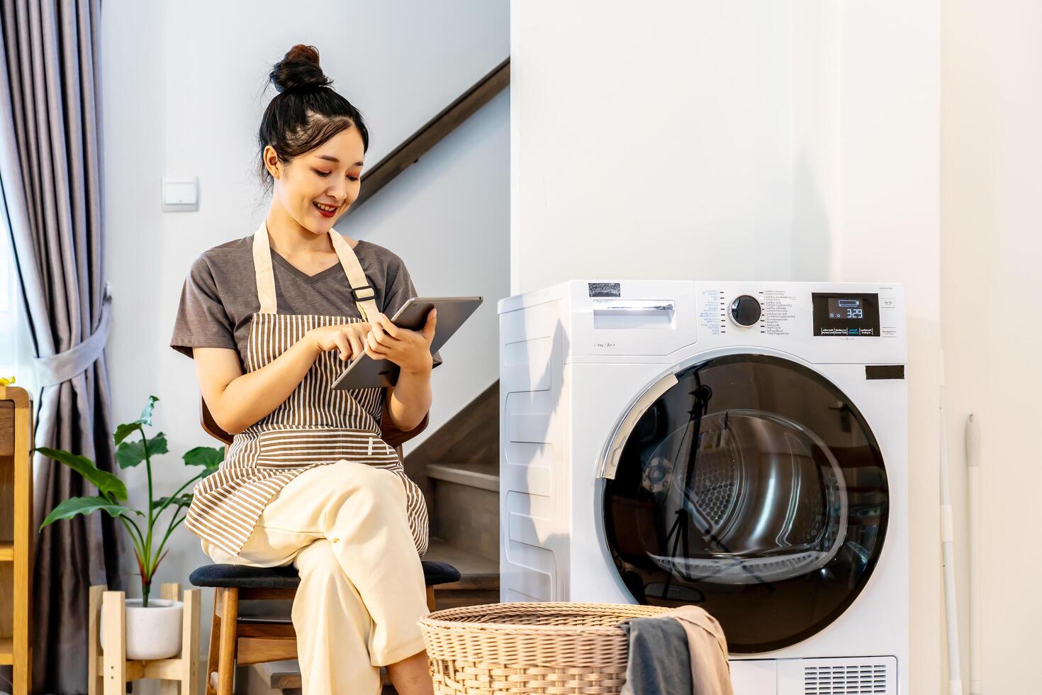 Young asian woman are playing with digital tablet in home sitting near the washing machine, while waiting for the washing machine to finish working photo