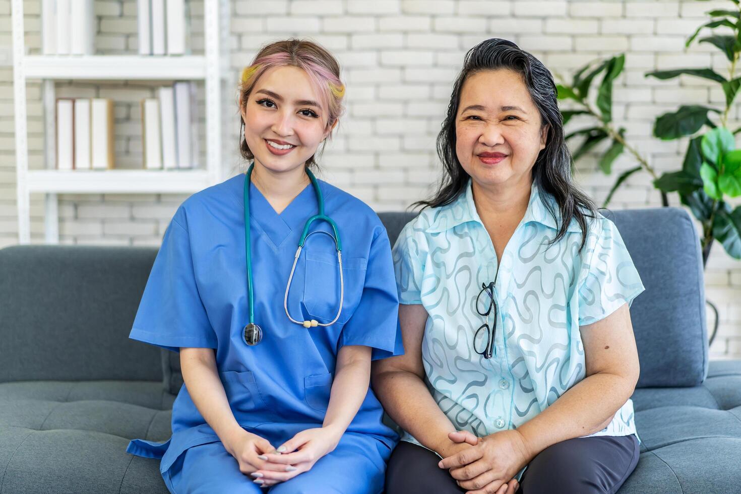 Portrait of smiling nursing home nurse in blue coat and older woman, Young friendly asian lady in medical uniform and senior female sitting on sofa and smiling at camera, Medical care concept photo