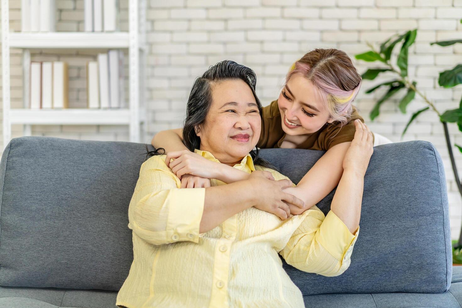 Happy asian young daughter embracing from back senior mother in law feeling respect and love, mature mom and adult daughter hugging and talking with looking at tv, spending leisure time together photo